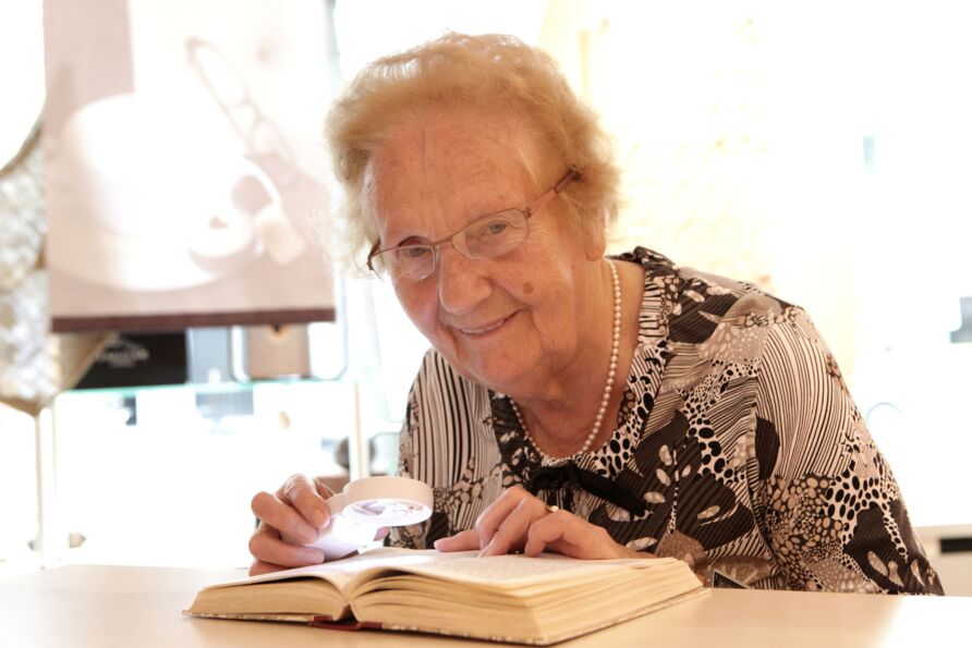 Foto: Seniorin mit rotblondem Haar und Brille sitzt an einem Tisch und lächelt den Betrachter an. In ihrer Hand eine Lupe mit Beleuchtung, darunter auf dem Tisch ein aufgeschlagenes Buch. Sie trägt eine Perlenkette und ein in Brauntönen gemustertes Kleid.