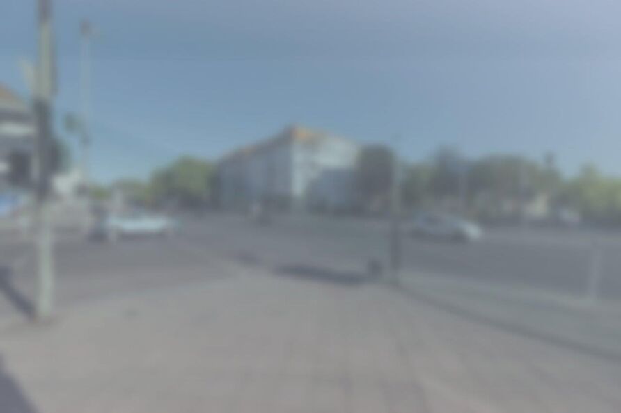 Foto: Blick über eine Kreuzung im Sonnenlicht – von einer Straßenecke zur diagonal gegenüberliegenden Ecke. Das Bild ist stark verschwommen und getrübt, die Kontraste sind reduziert. Zwei Autos sind zu erkennen.