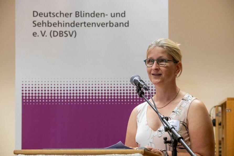 Foto: Christiane Möller spricht in ein Mikrofon. Sie ist Anfang 40, trägt ein helles, sommerliches Kleid und eine Brille. Die blonden Haare sind am Hinterkopf zusammengesteckt. Im Hintergrund ein Roll-up des Deutschen Blinden- und Sehbehindertenverbands.