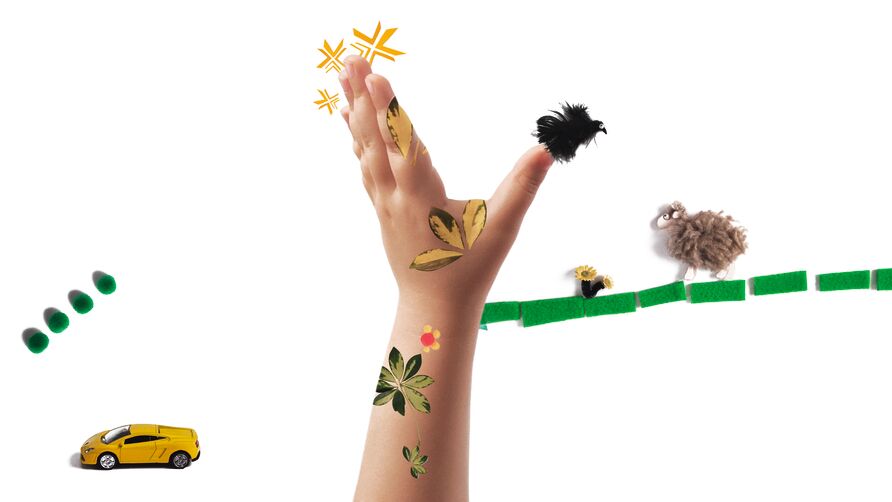Eine Kinderhand ragt von unten in die Bildmitte. Auf der Haut sind bunte Pflanzenmotive zu sehen, drumherum sind Tastbuchmaterialien arrangiert, darunter ein Spielzeugauto, ein Schaf und ein Vogel.