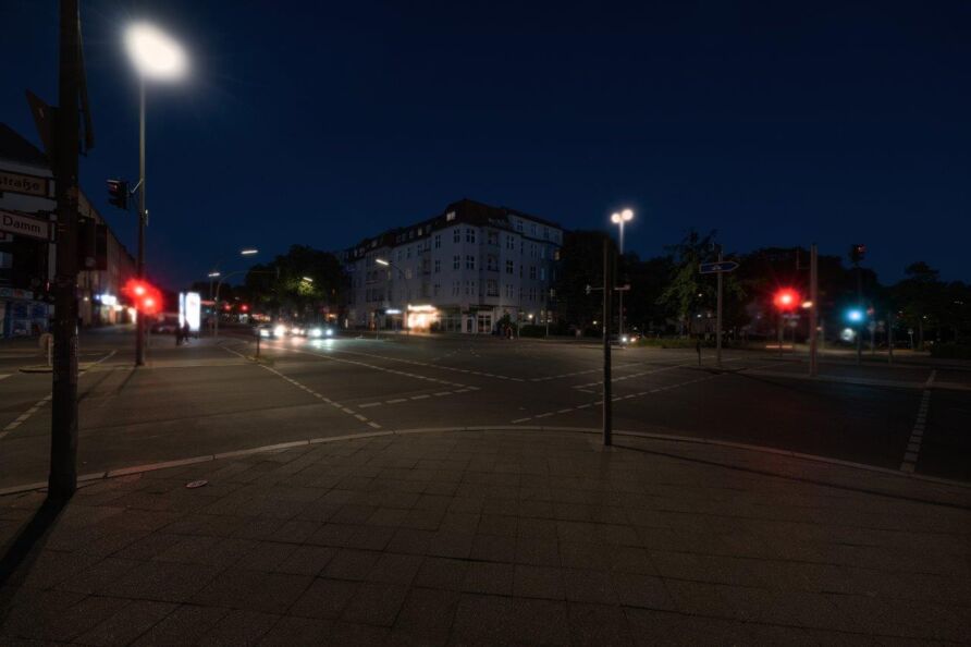 Foto: Blick über eine Kreuzung bei Nacht – von einer Straßenecke zur diagonal gegenüberliegenden Ecke. Die Straßen sind mehrspurig mit Autos und zu Fuß Gehenden. Das Bild ist getrübt, die Lichtquellen in verschiedenen Farben sind gedämpft.
