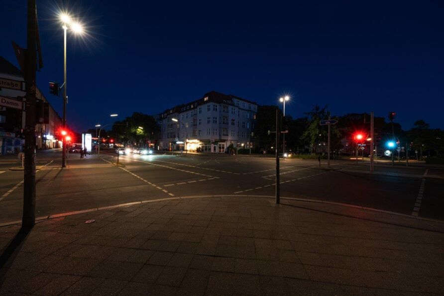 Foto: Blick über eine Kreuzung bei Nacht – von einer Straßenecke zur diagonal gegenüberliegenden Ecke. Die Straßen sind mehrspurig mit Autos, zu Fuß Gehenden und Lichtquellen in verschiedenen Farben (Ampeln, Straßenlampen, Autoscheinwerfer …).