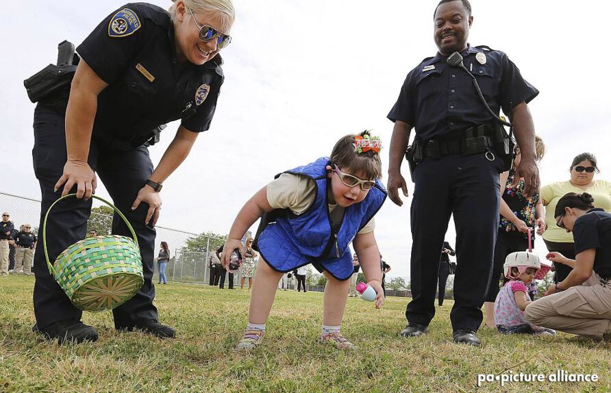 Ein sechsjähriges Mädchen steht mit konzentriertem Gesichtsausdruck gebückt auf einer Wiese, in beiden Händen je ein Kunststoffei. Links von ihr steht eine lächelnde Frau mit einem Korb, rechts ein lächelnder Mann, beide in amerikanischer Polizeiuniform.